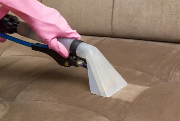 A hand wearing a pink rubber glove vacuums a brown sofa with a distinct difference between the clean and dirty parts of the seating area