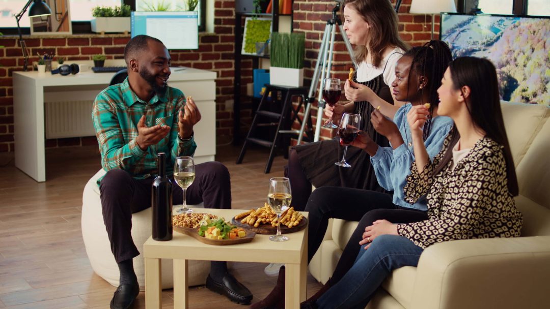 three young adults sat on a sofa drinking wine and food on the coffee table in front of them and a fourth friend sat opposite